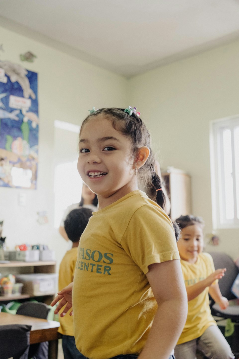 Young girl in a yellow PASOS CENTER shirt smiles in a classroom.